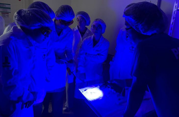 Acadêmicos da UFMS durante demonstração no laboratório, sob penumbra e luz totalmente azul. Tal item é utilizado para identificar elementos comprobatórios em crimes.