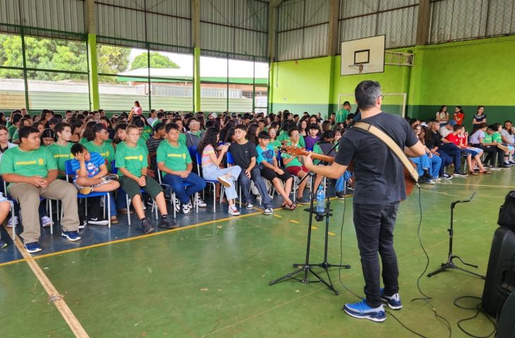 Professor com violão em uma quadra escolar coberta tocando alguma música para centenas de alunos, sentados em cadeiras, atentos a ele.