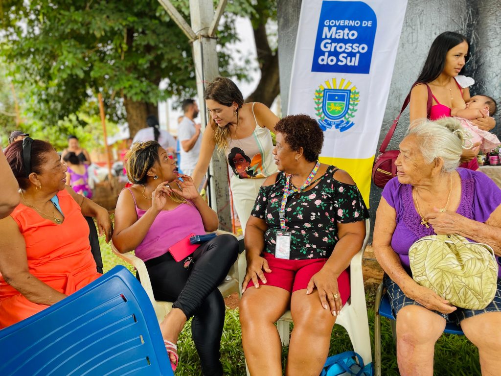 Mulheres  com roupas coloridas são recebiddas pela secretária Viviane Luíza, que usa um vestido cumprido que estampa a figura de uma mulher.