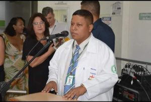 Presidente do Conselho Comunitário de Segurança do Parati, Irwing Ferreira, está de gravata azul e camisa branca em frente a um microfone.