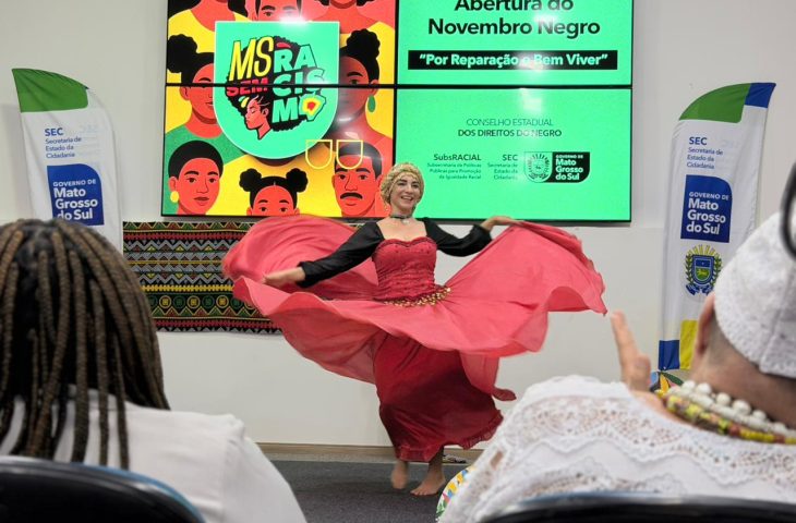 Feature image Mulher sorridente com uma saia longa e vermelha rodada realiza uma performance de dança no palco. Duas pessoas do público vestidas de branco são vistas em primeiro plano