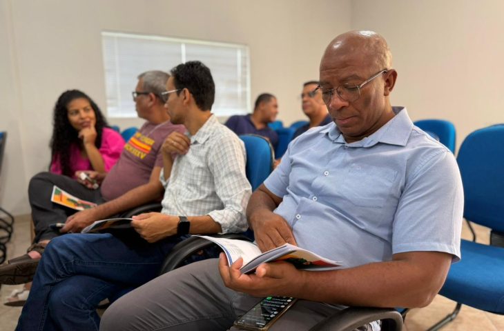 Um homem negro de meia-idade, vestindo uma camisa social azul clara e óculos, está sentado em uma cadeira azul e foca a atenção na leitura do guia MS sem Racismo. Outras pessoas estão sentadas ao fundo, algumas interagindo.