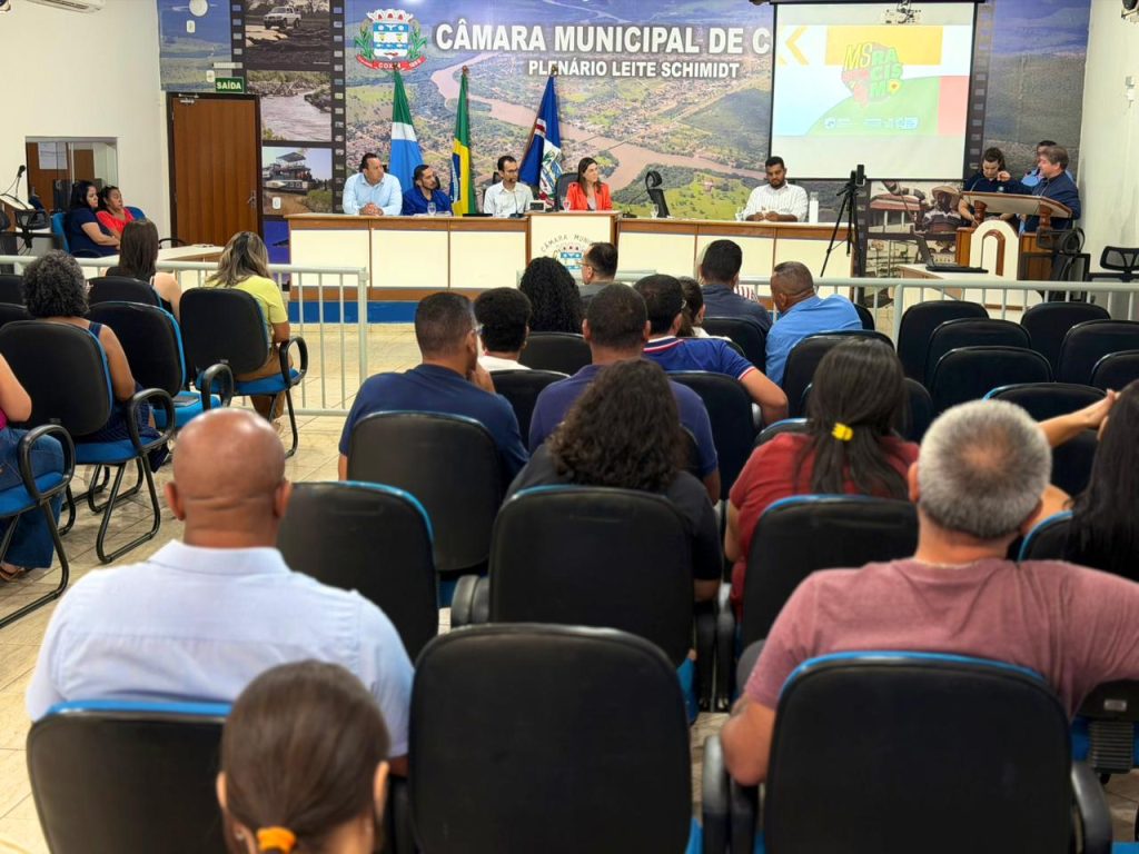 Vista geral do Plenário Leite Schmidt da Câmara Municipal de Coxim, durante um evento. Na mesa diretora, há um grupo de pessoas discursando ou participando, enquanto a plateia lotada assiste de frente, vista predominantemente por trás (nuca).
