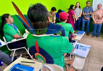 Vista de um participante indígena, de costas, sentado em uma carteira de sala de aula. Ele usa uma camiseta verde e um cocar grande com penas azuis, vermelhas e pretas. Na frente dele há um caderno "Empretec". Outros participantes e instrutores estão em pé ao fundo.