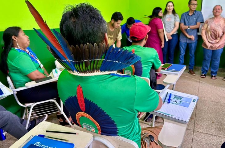 Vista de um participante indígena, de costas, sentado em uma carteira de sala de aula. Ele usa uma camiseta verde e um cocar grande com penas azuis, vermelhas e pretas. Na frente dele há um caderno "Empretec". Outros participantes e instrutores estão em pé ao fundo.